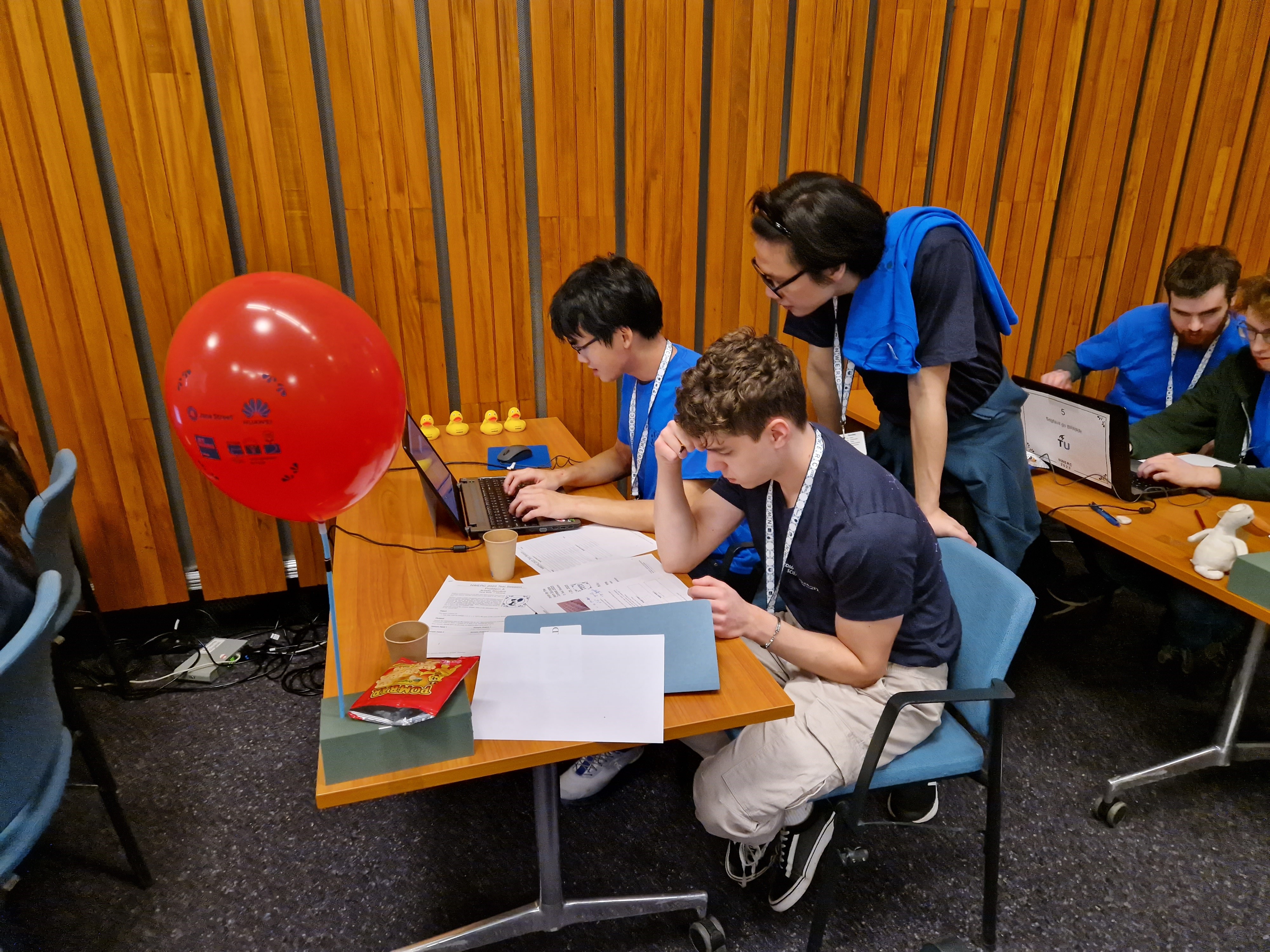 Three computer programming students from Team NewJeans (two seated, one standing) solving computer programming problems on a laptop. There is paper on the desk, an empty crisp packed, a row of yellow ducks and a red balloon - indicating they've solved one puzzle already&nbsp;&nbsp;