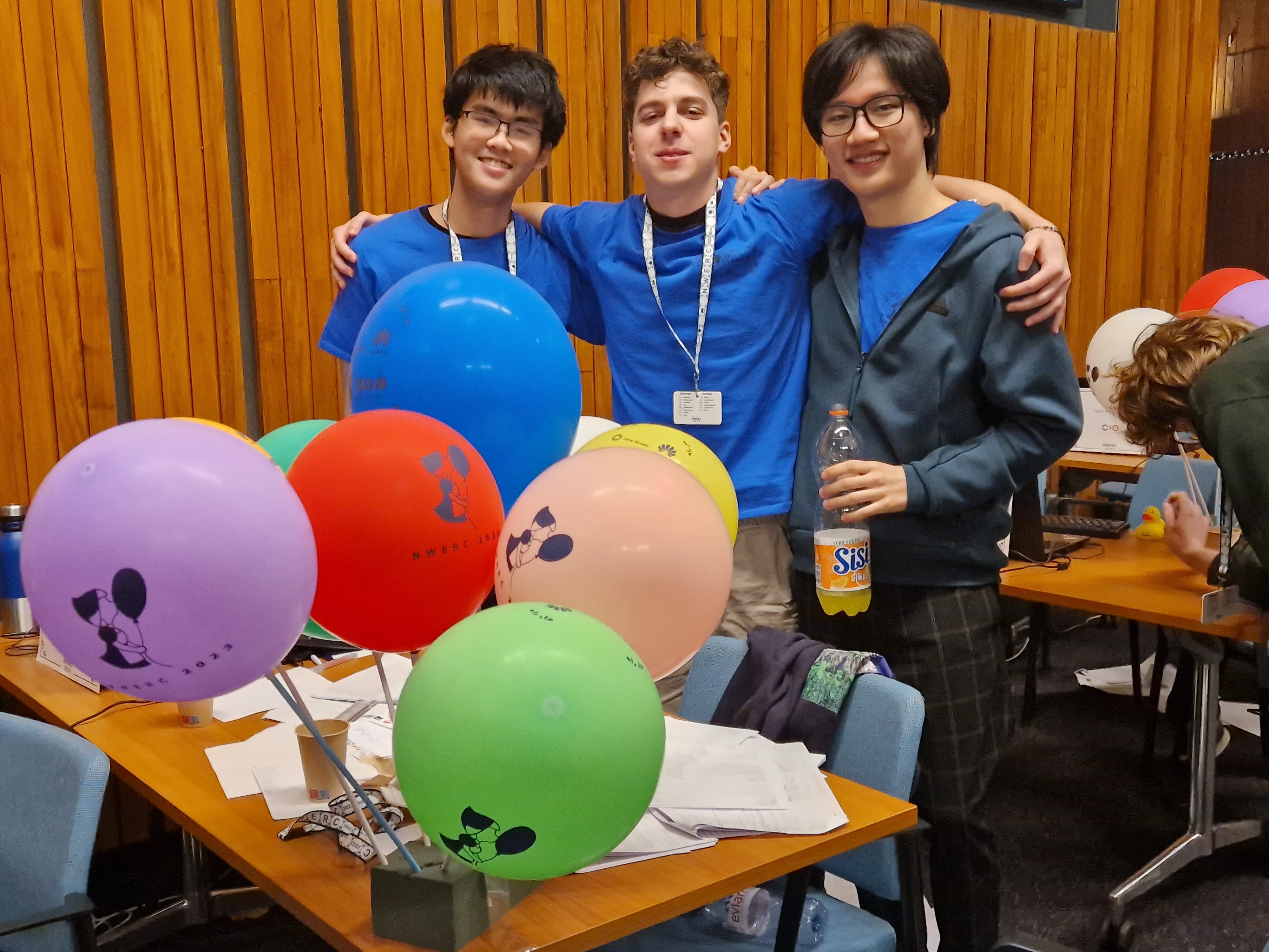 Three members of Team NewJeans smile after they win the contest, surrounded by colourful ballons