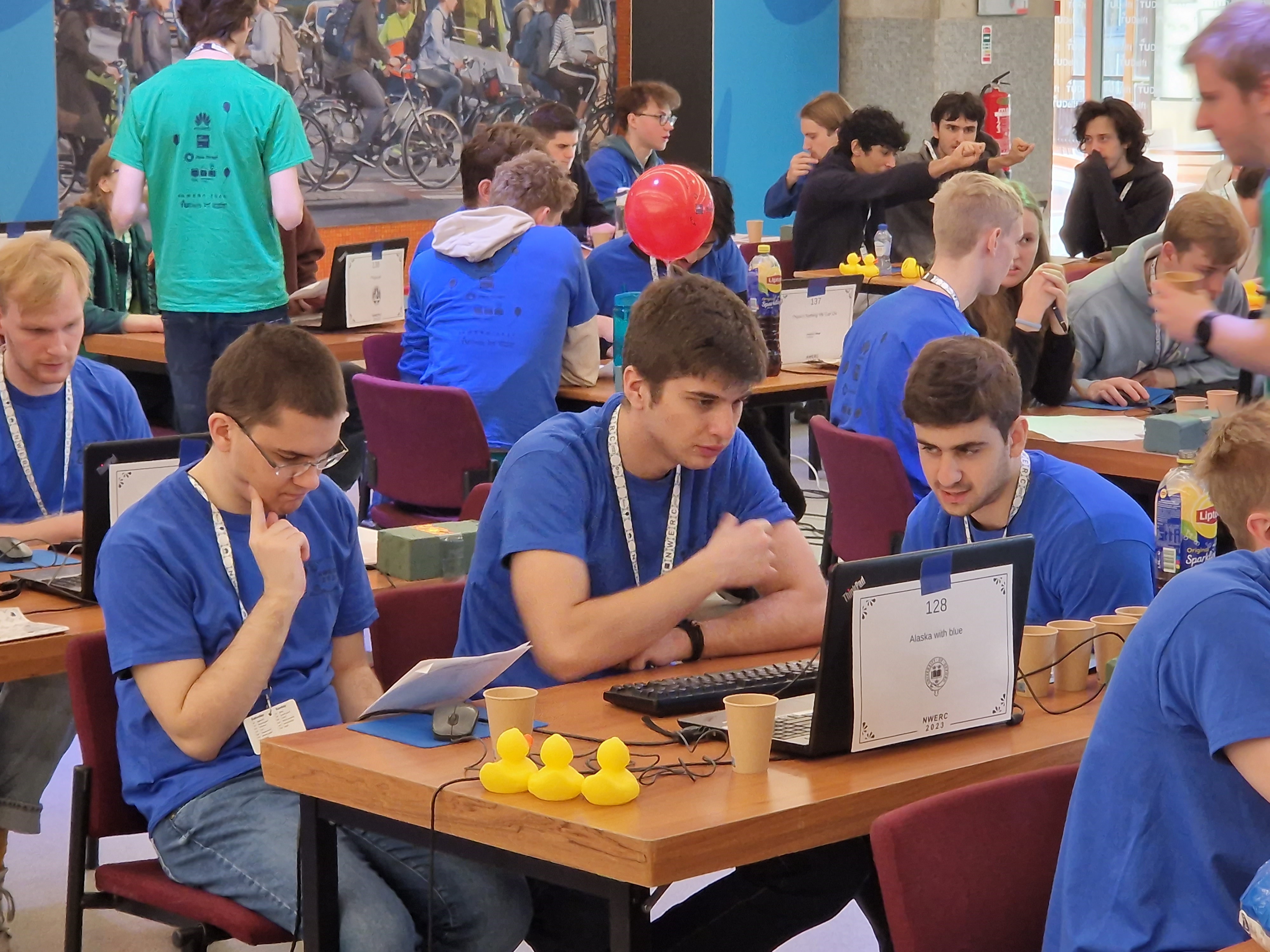 Team Alaska with blue during the competition. The three team members are in a busy room, seated and working around one laptop computer. There are papers and yellow ducks on the desk.