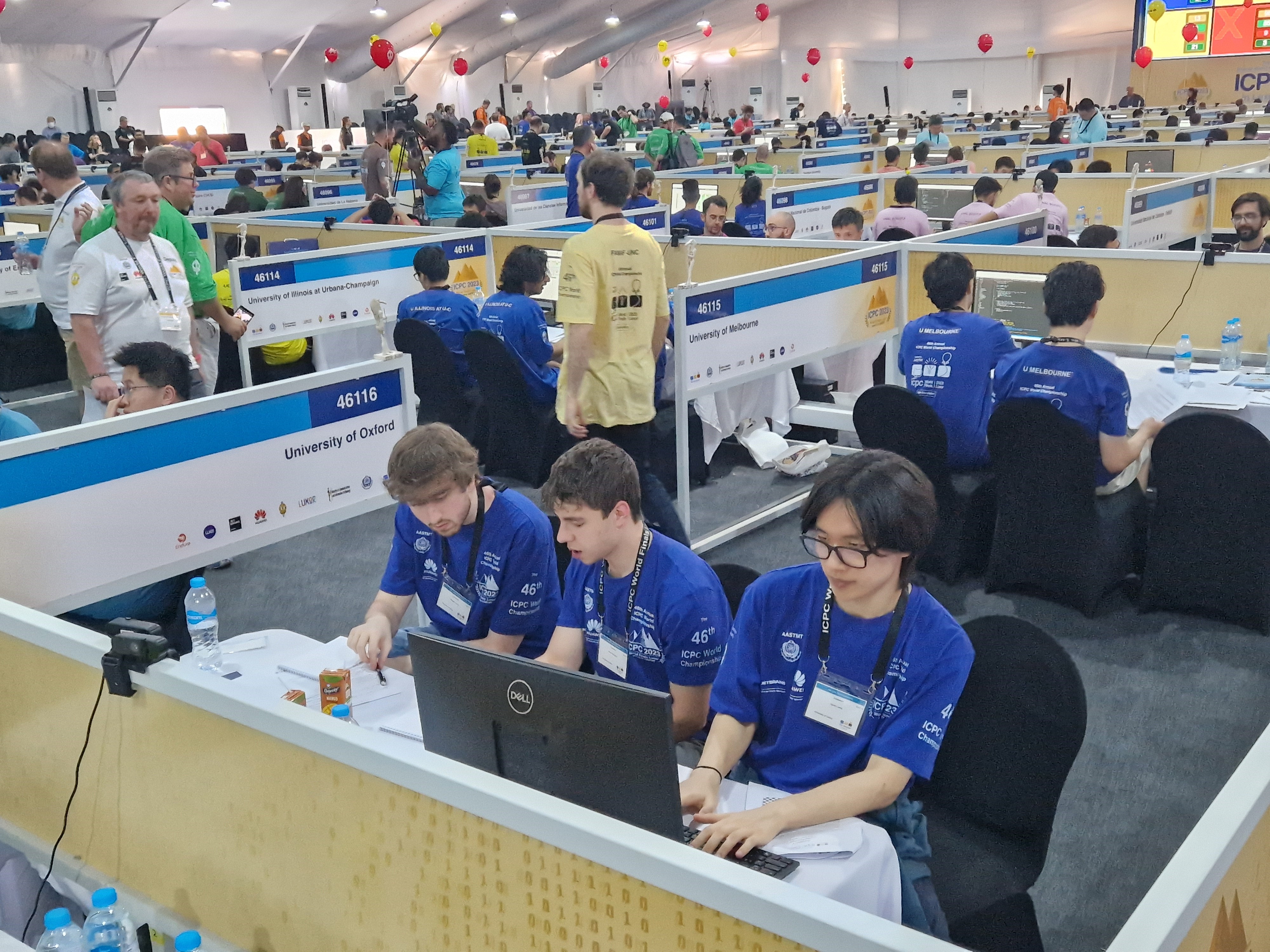 A photograph of three people sat to a desk whilst wearing blue tops and landyards. they are in a room with lots of booths with computers, all the other booths in the background are filled with people.