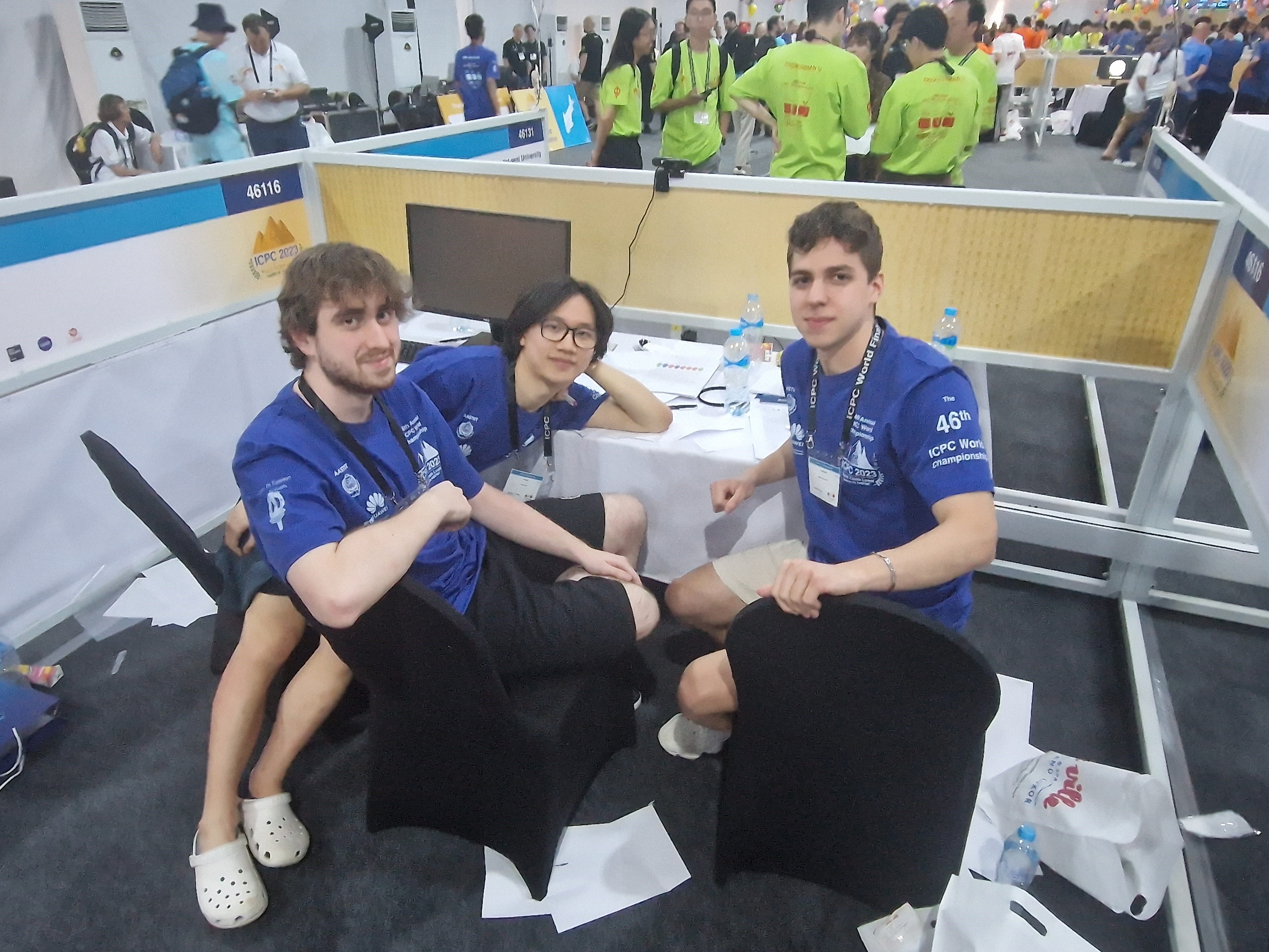 a photograph of three people looking at the camera, sat at a desk. They are wearing blue tops and landyards and are sat on black chairs. They are in a large room filled with people in the background.