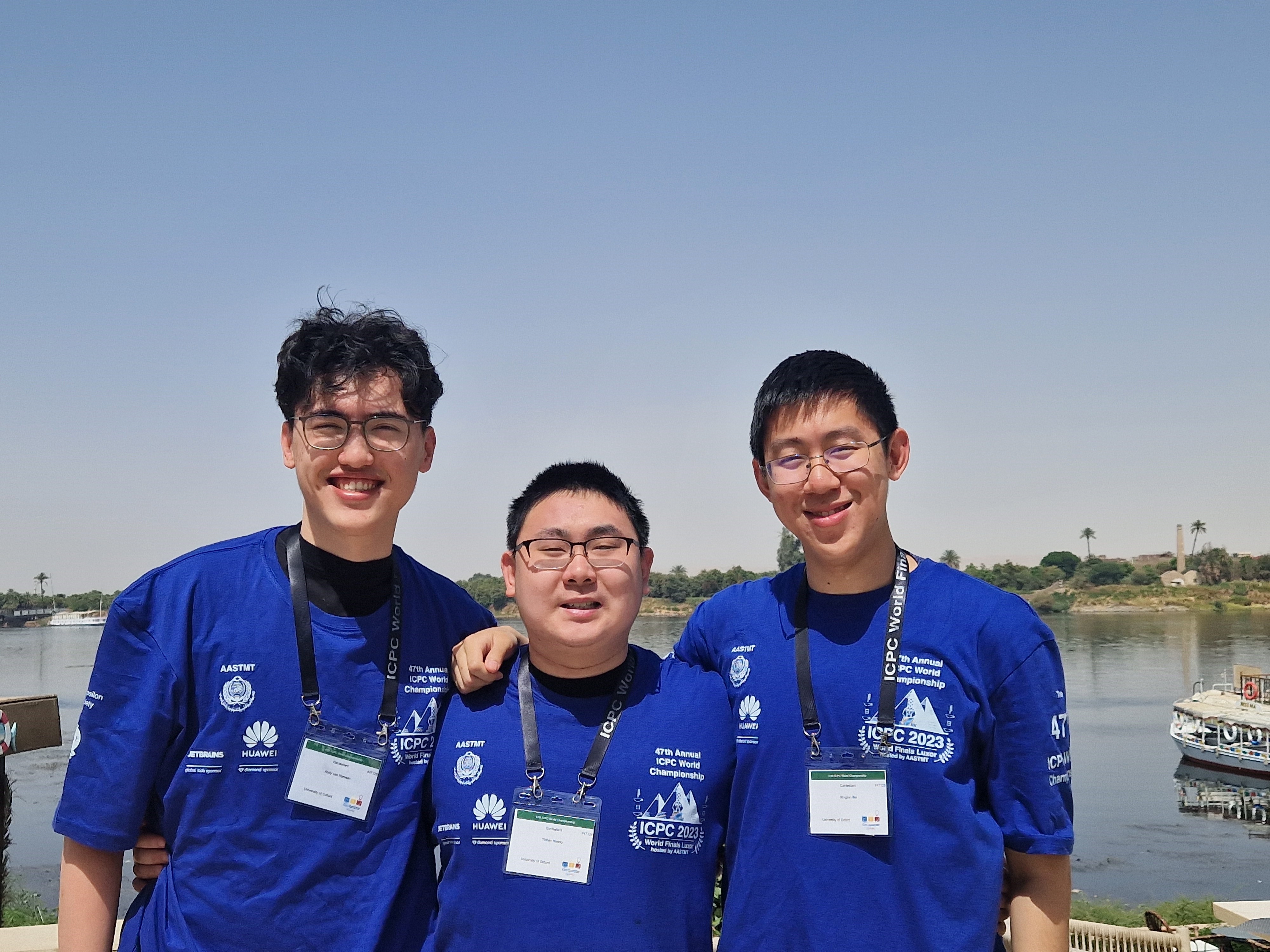 A photograph of three people looking at the camera whilst wearing blue tops and landyards. They are stood outside near water, under a hazy blue sky.