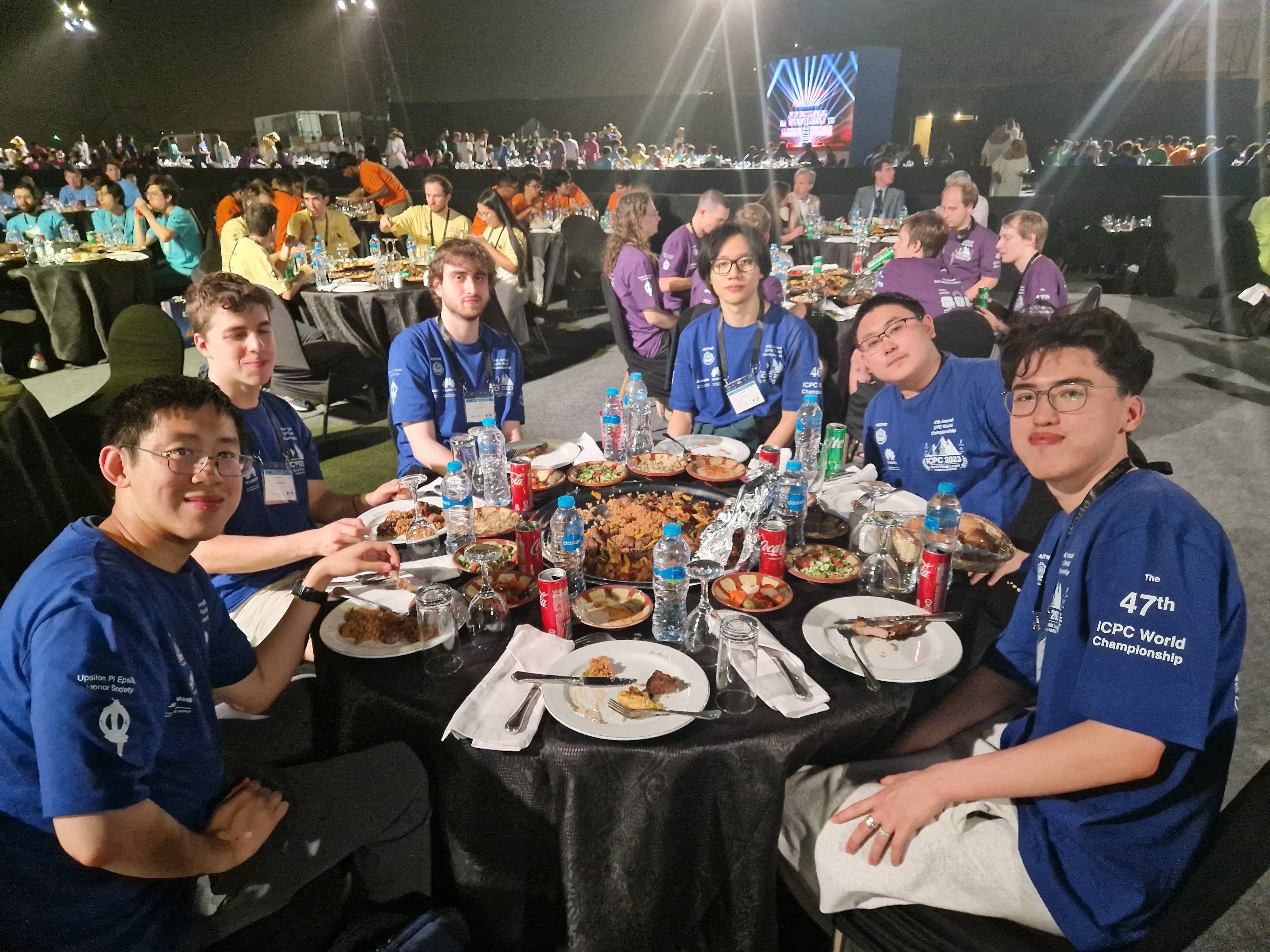 A photograph of six people sat at a round table, all looking at the camera. They are wearing blue tops and have plates of food in front of them. they are sitting in a dark room with other tables with people sat at them.