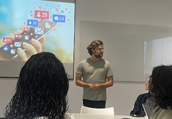 A photograph of Research Associate Ulrik Lyngs stood at the front of a room by a projected screen.