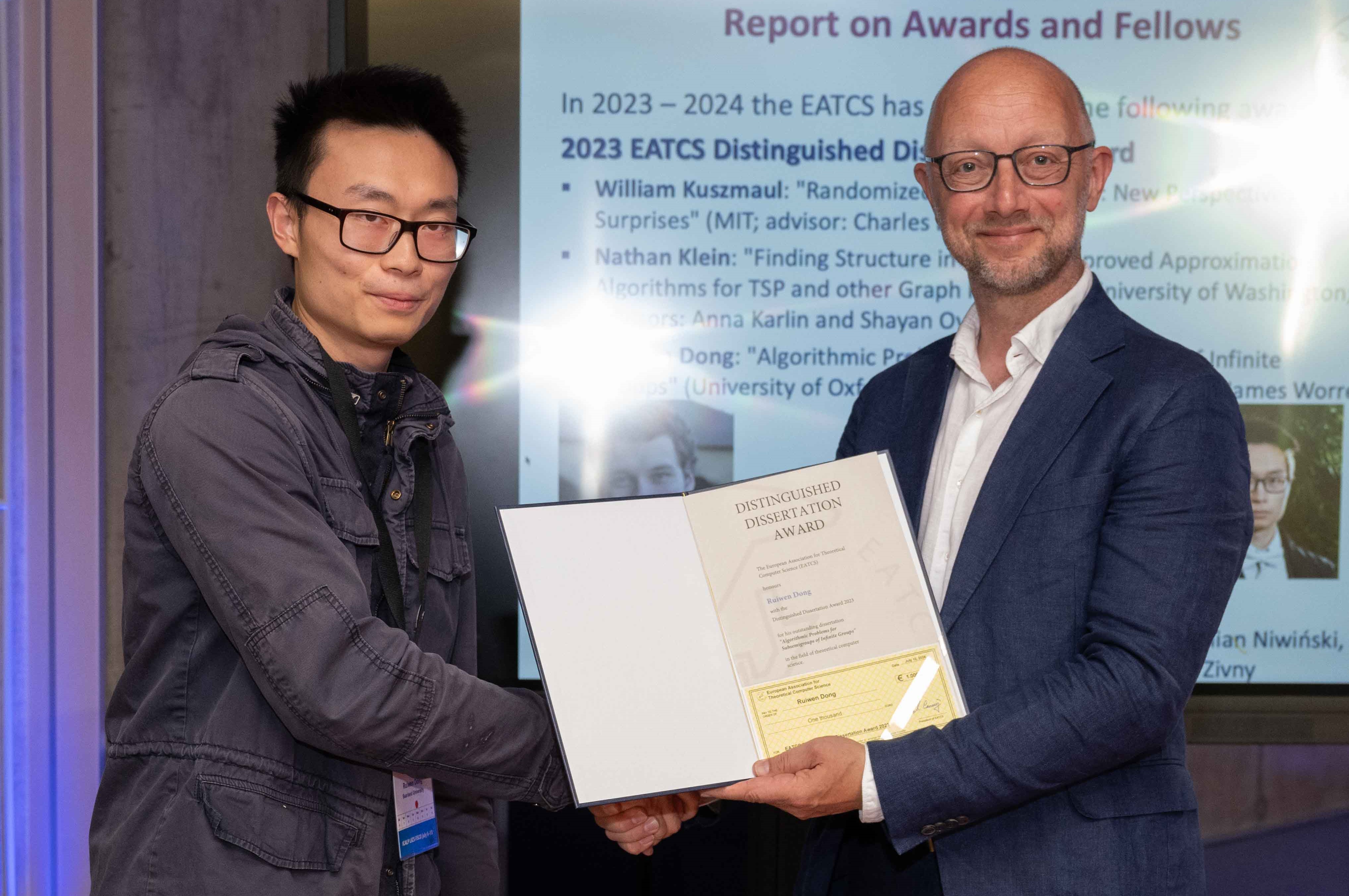 Image of Ruiwen Dong receiving his Distinguished Dissertation Award award at the ICALP Conference. He is shaking hands with an unmamed man who is displaying the certificate award.