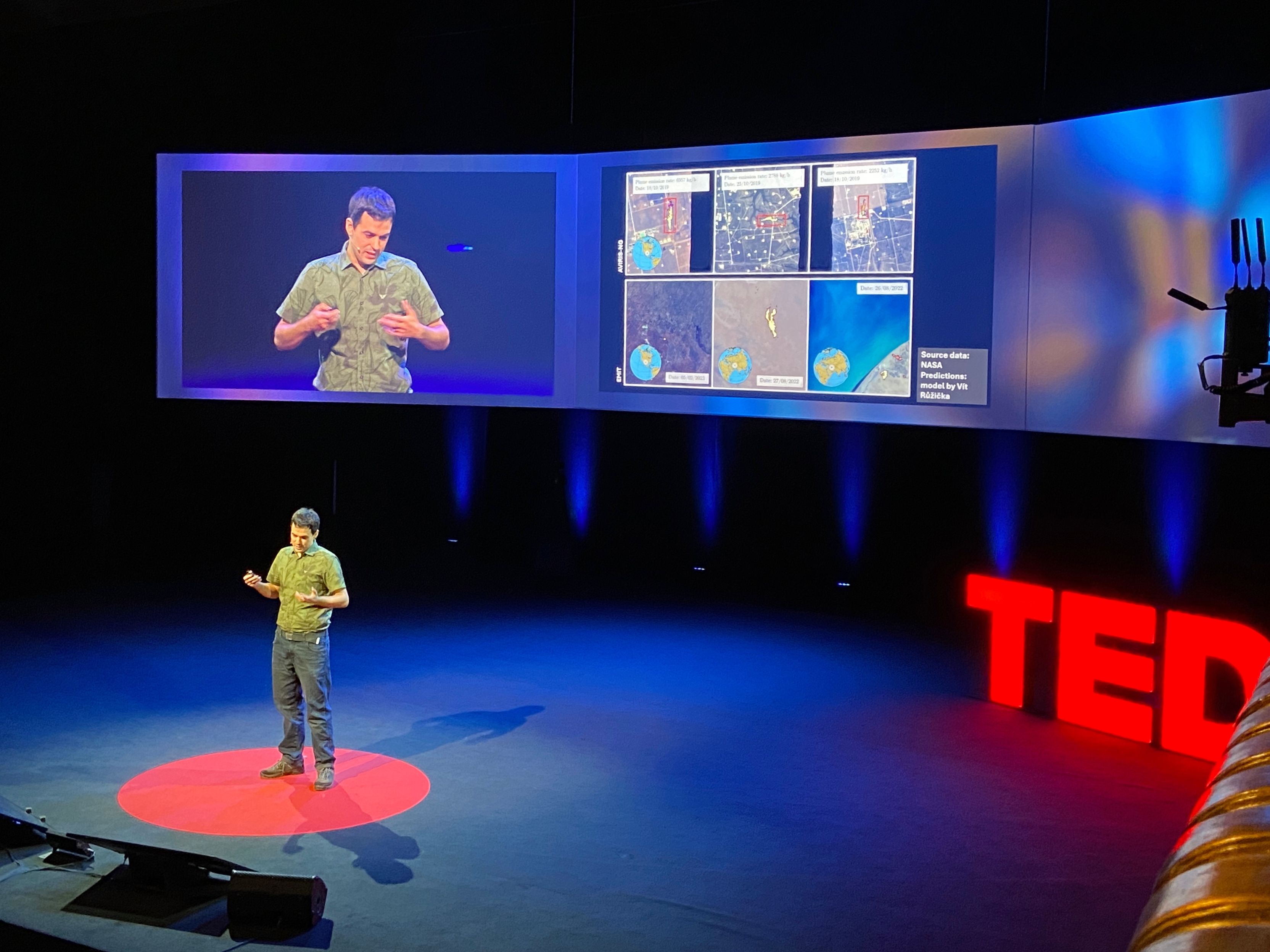 A photograph of a man in a green top standing on a red circle on a dark stage. Behind the man there is a series of large screens displaying an image of himself of stage and graphics. At the back right hand side of the stage there is a red light TED sign.